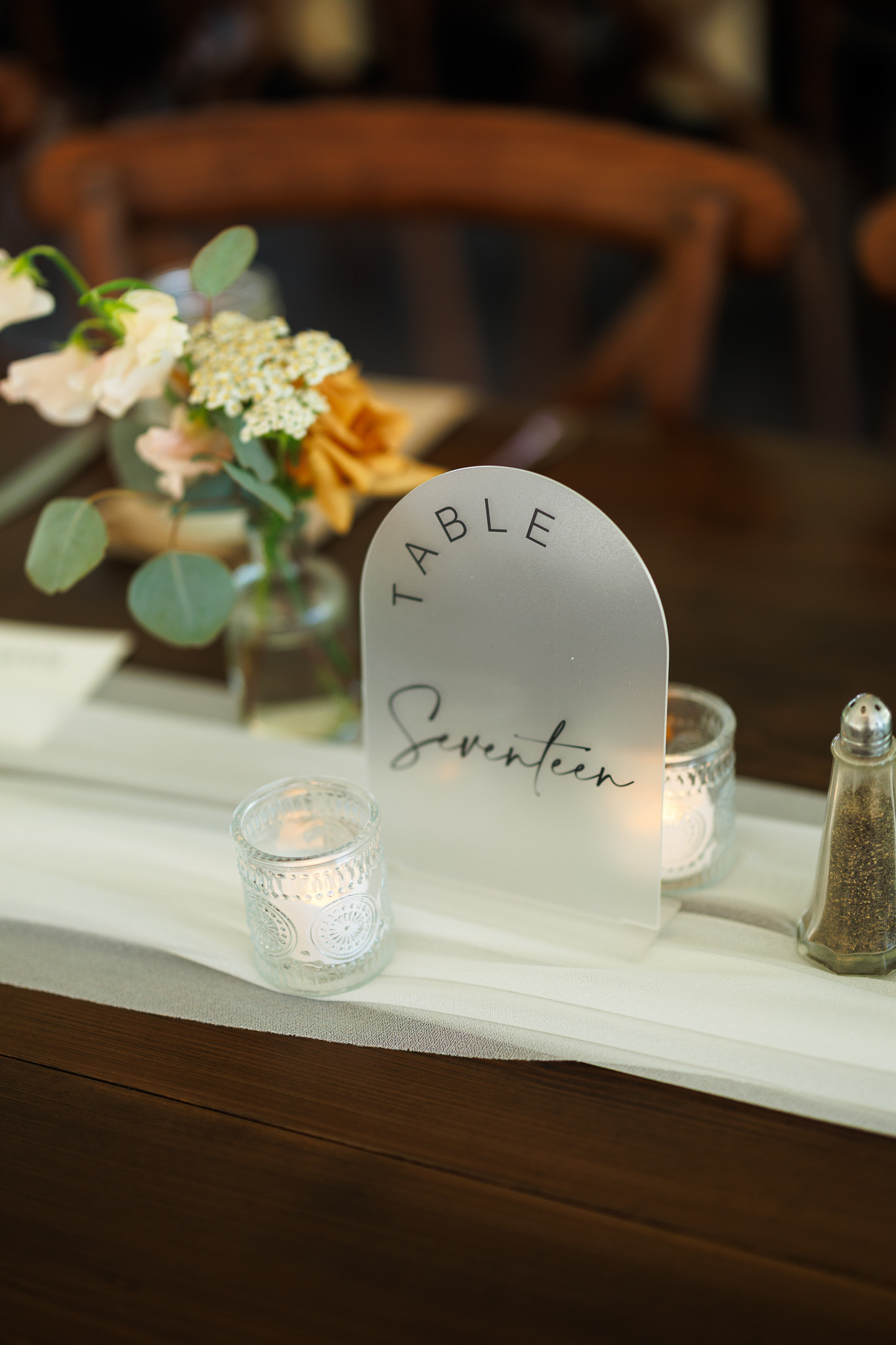 Modern Wedding Table Number with Floral Centerpiece Acrylic Table Seventeen sign styled with candlelight and a small arrangement of greenery and warm neutral flowers on a wooden reception table.