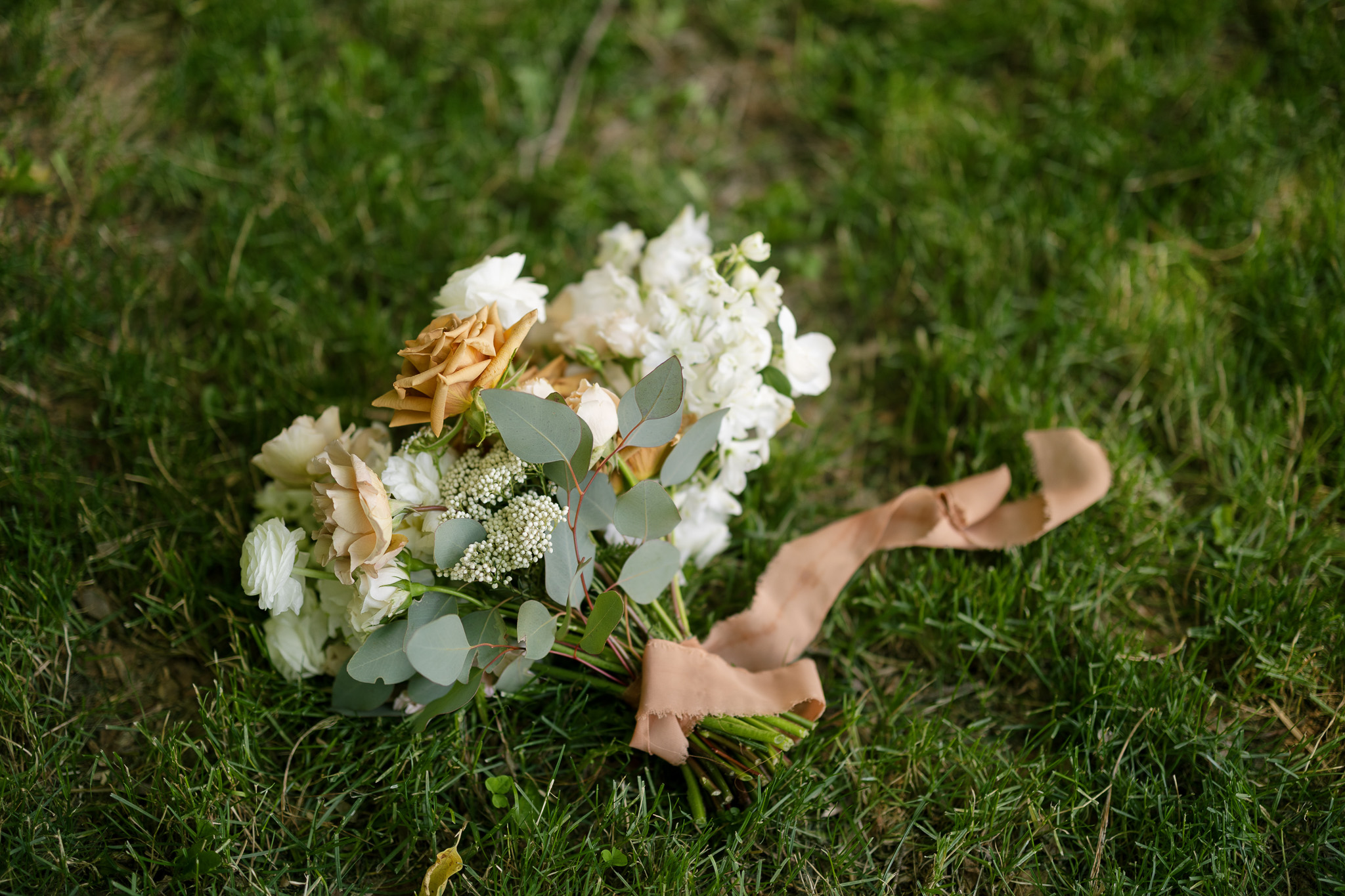 Neutral Wedding Bouquet with Eucalyptus Bridal bouquet featuring white and warm neutral flowers, eucalyptus greenery and a soft tan ribbon resting on the grass.