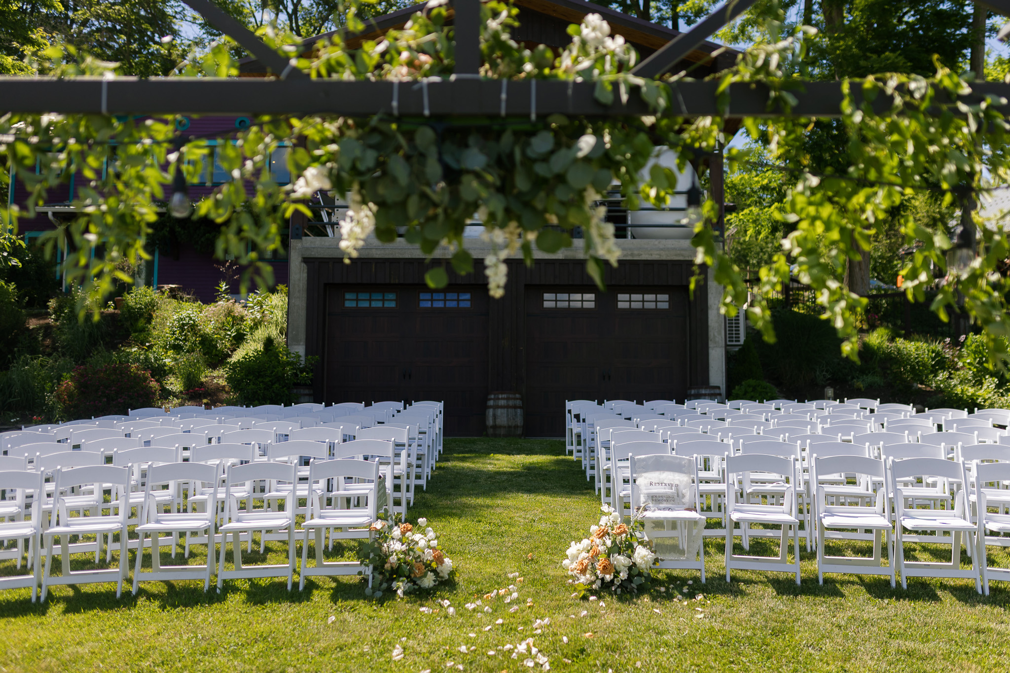 Outdoor Garden Wedding Ceremony Setup Outdoor wedding ceremony setup with rows of white chairs, aisle florals, scattered petals and greenery decorating the ceremony structure.
