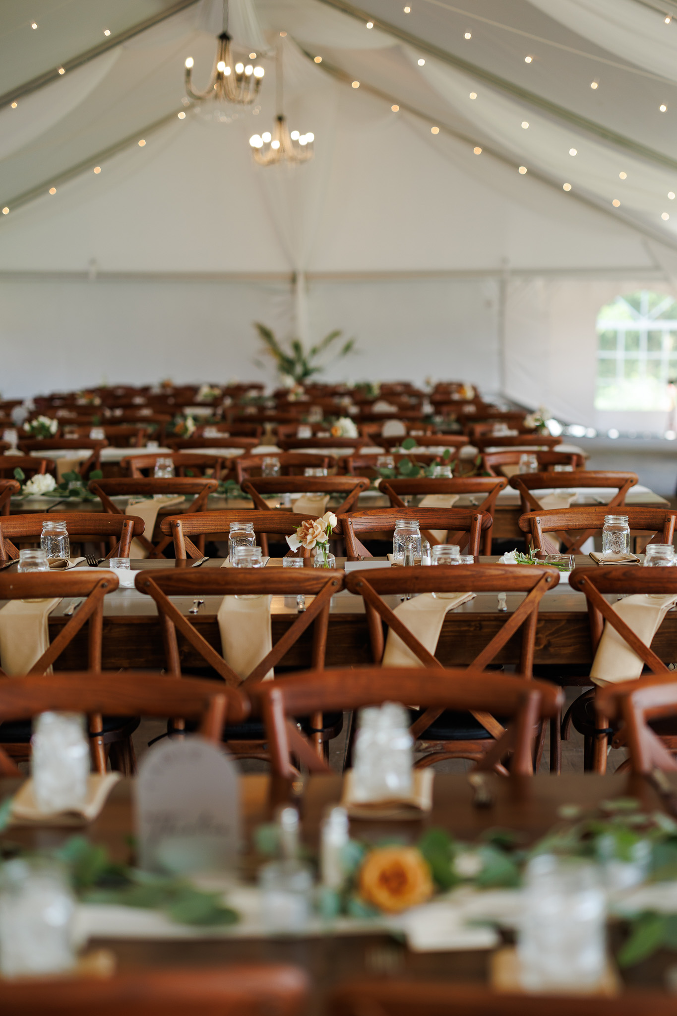 Outdoor Tent Wedding Reception with Farm Tables Wedding reception inside a white sailcloth tent with long wooden farm tables, cross-back chairs, greenery, neutral linens, chandeliers and string lights.