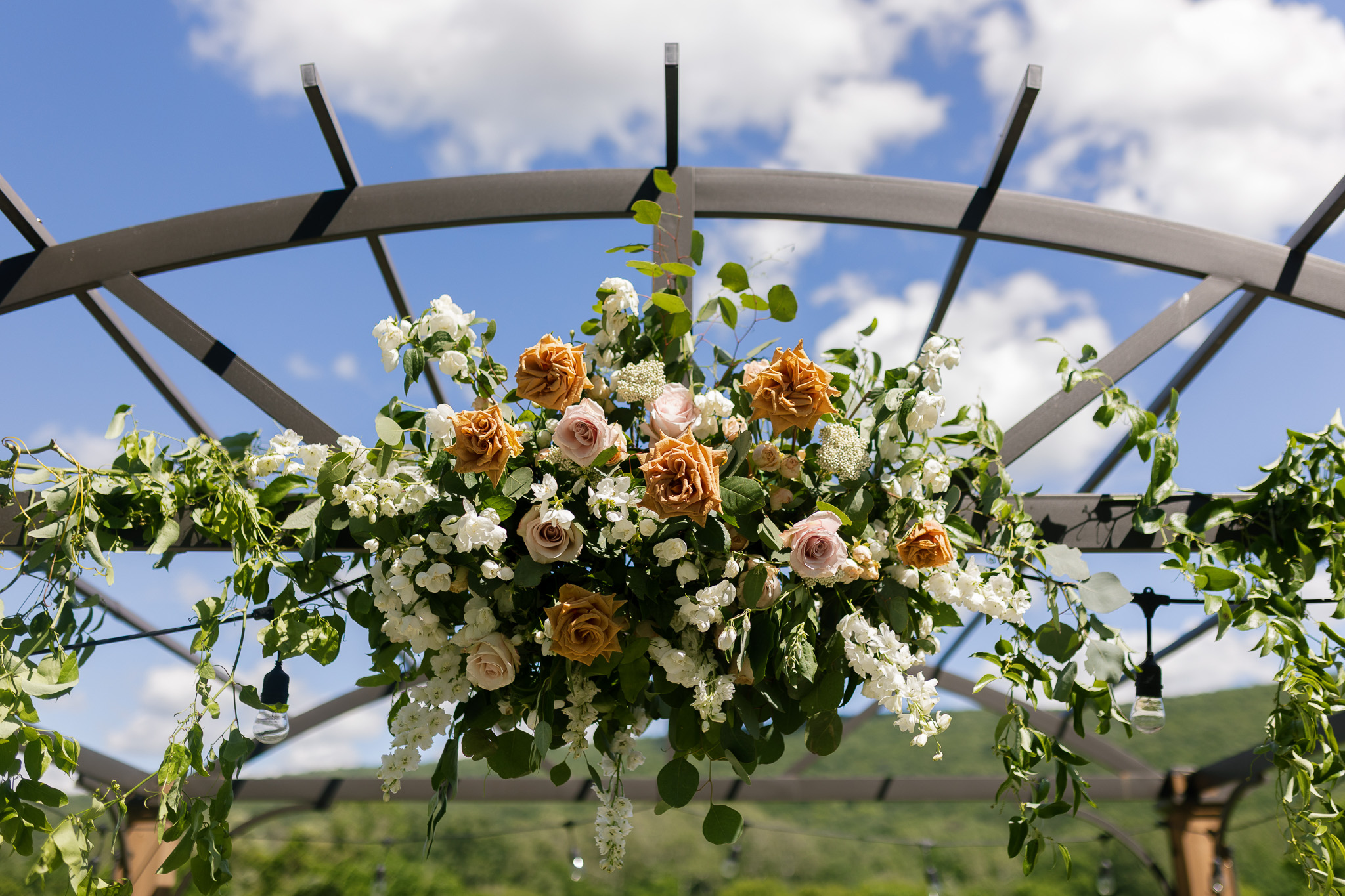 Romantic Wedding Ceremony Floral Installation Large ceremony floral installation featuring warm caramel and blush roses, white flowers and abundant greenery against a blue summer sky.