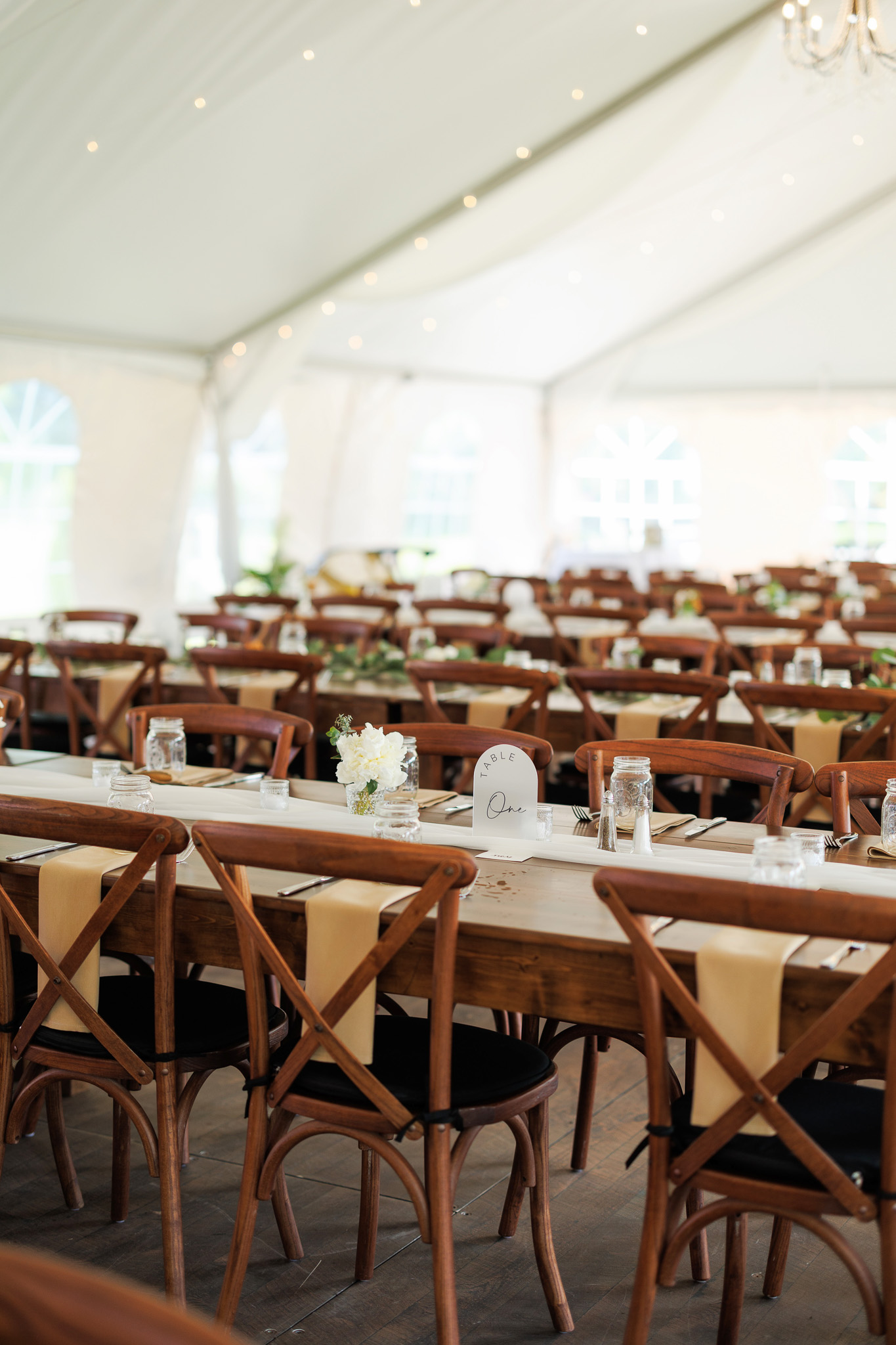 Rustic Wedding Reception Table Setup Long wooden reception tables arranged with cross-back chairs, neutral runners, mason jars and simple floral centerpieces inside a bright wedding tent.