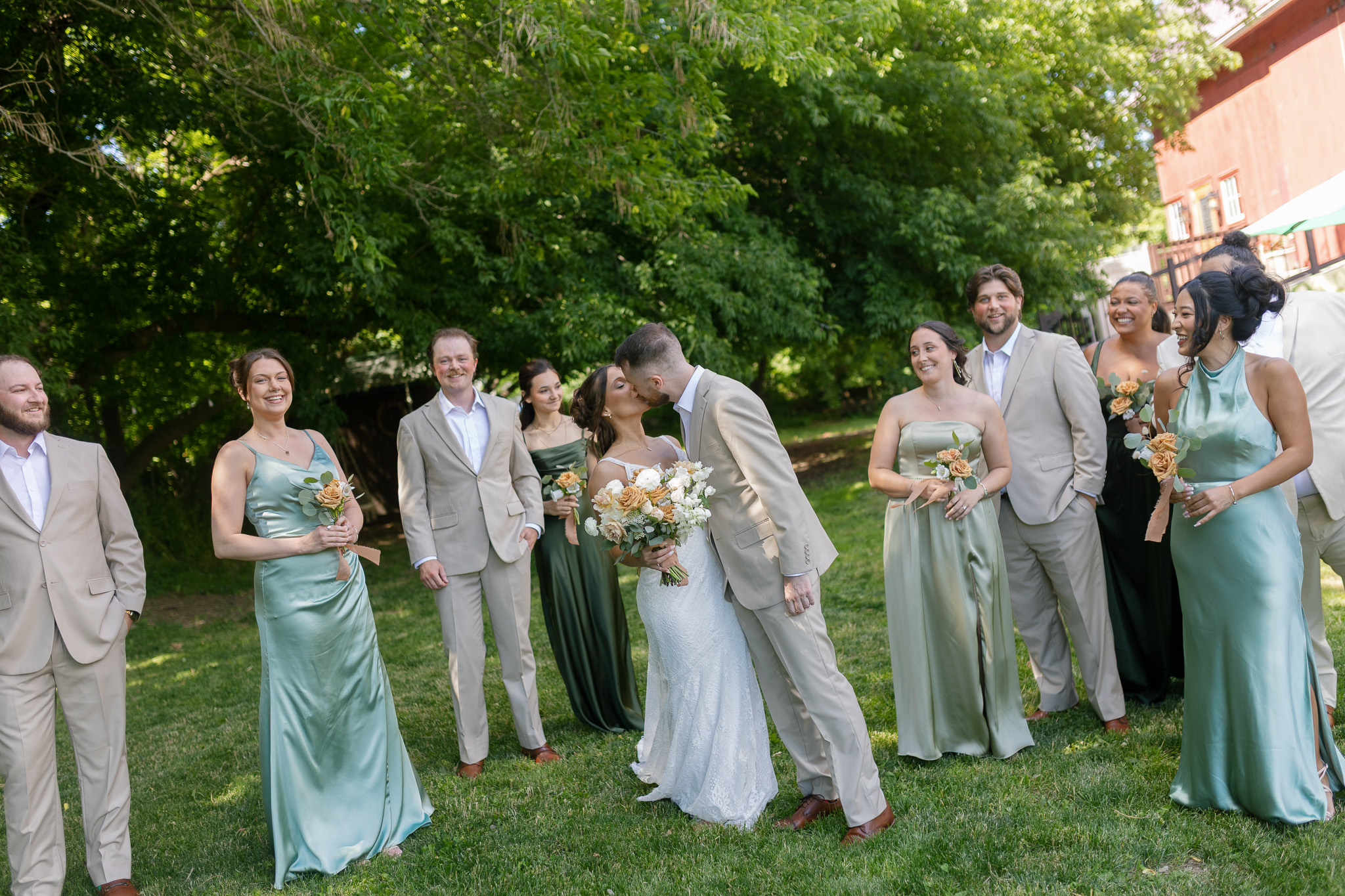 Sage Green and Neutral Wedding Party Bride and groom kissing surrounded by their wedding party dressed in sage green, dark green and light neutral wedding attire.