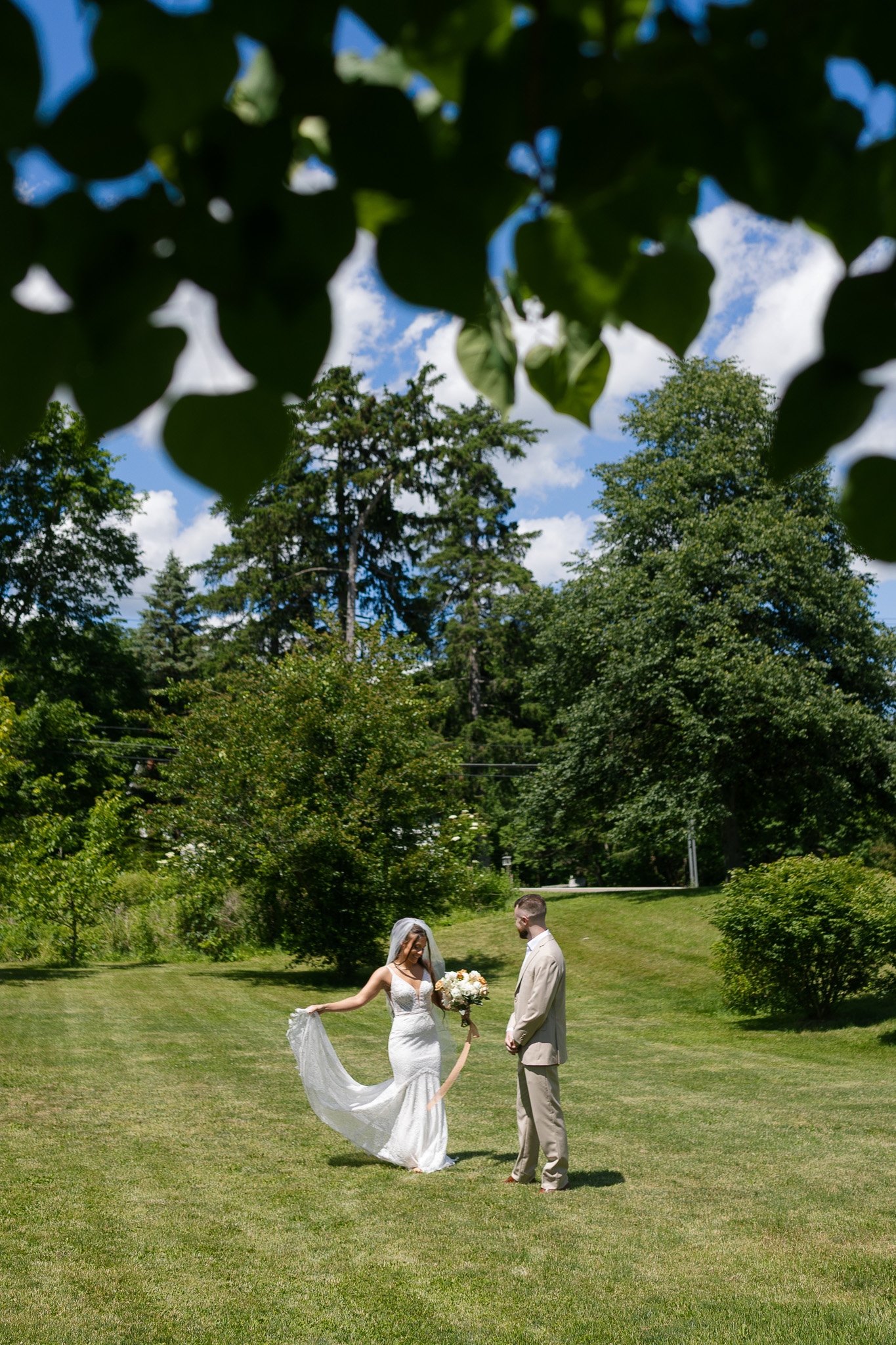 Summer Outdoor Wedding Couple Portrait Bride holding her train and bouquet while standing with the groom on a green lawn beneath leafy trees and a blue summer sky.
